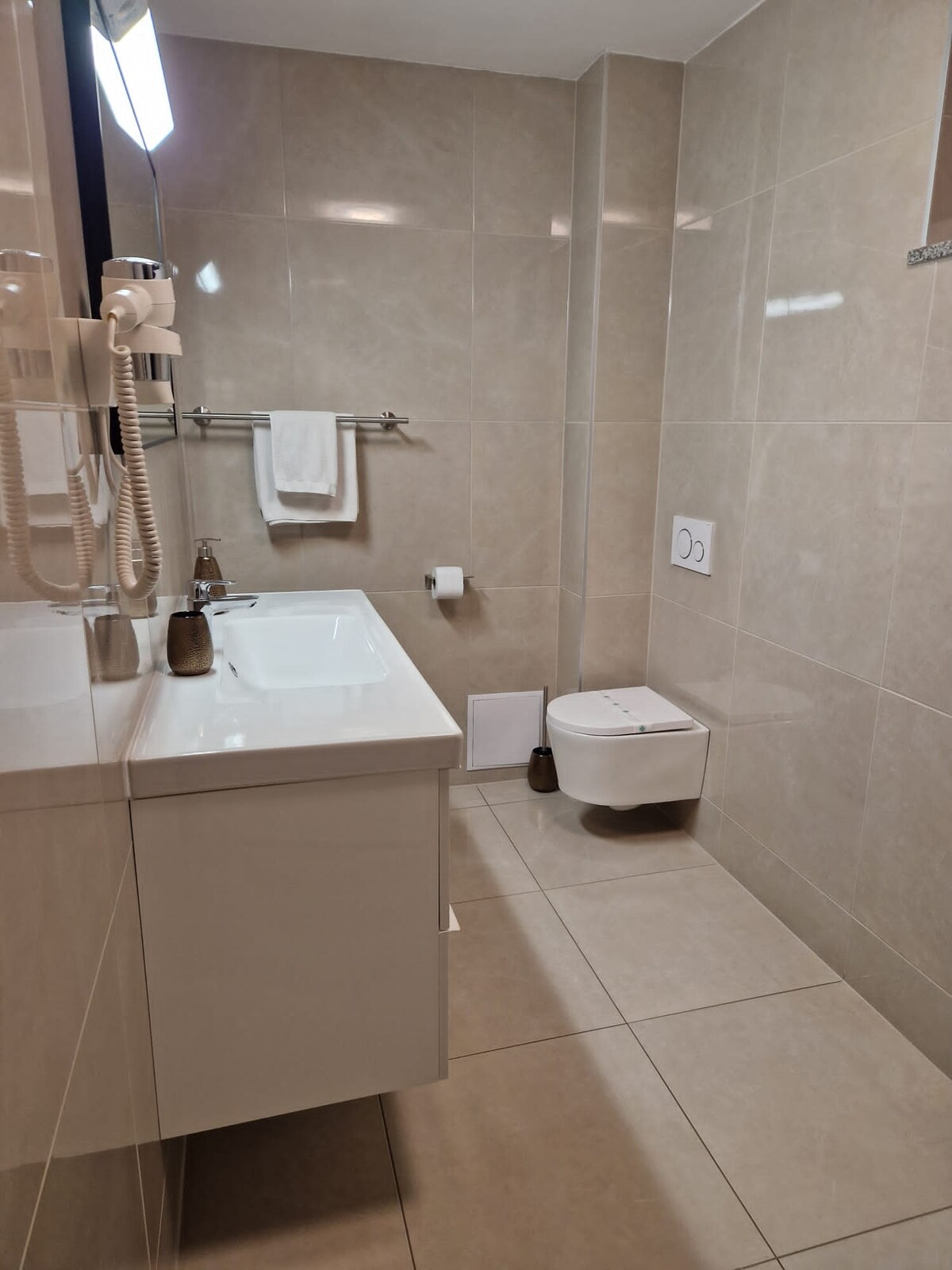 Cream tiled bathroom with a white vanity unit and basin, a mirror with a hairdryer beside it, a towel rail with a white towel and a wall-hung toilet on the right