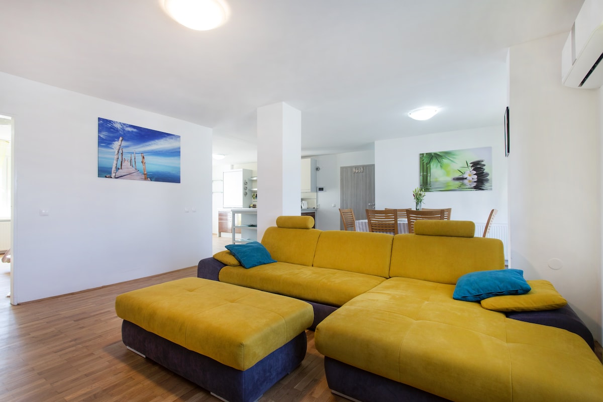 A large mustard-yellow corner sofa with teal cushions and a matching footstool on a wooden floor, with the kitchen and a dining table with wooden chairs visible beyond