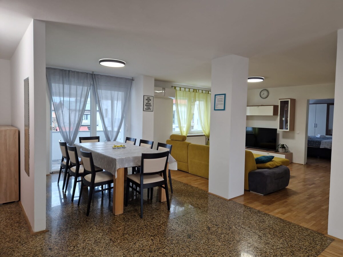 Veliki otvoreni prostor apartmana br. 2: dugi blagovaonski stol na kamenom podu uz gorušičastu kutnu garnituru i televizor