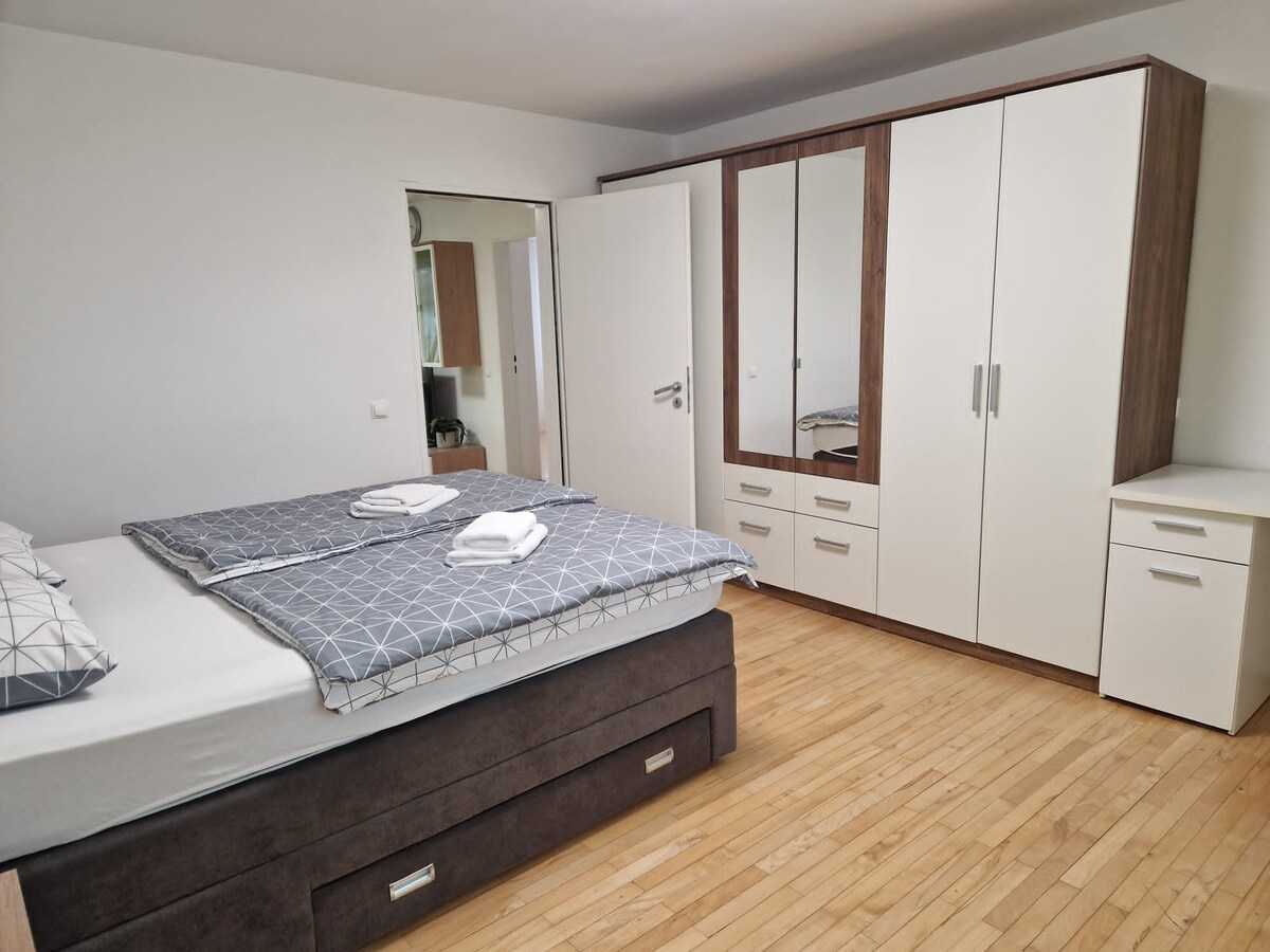 Bedroom seen from the bed towards a tall white and walnut wardrobe with drawers and a mirrored panel, a small chest of drawers beside it and a wooden floor