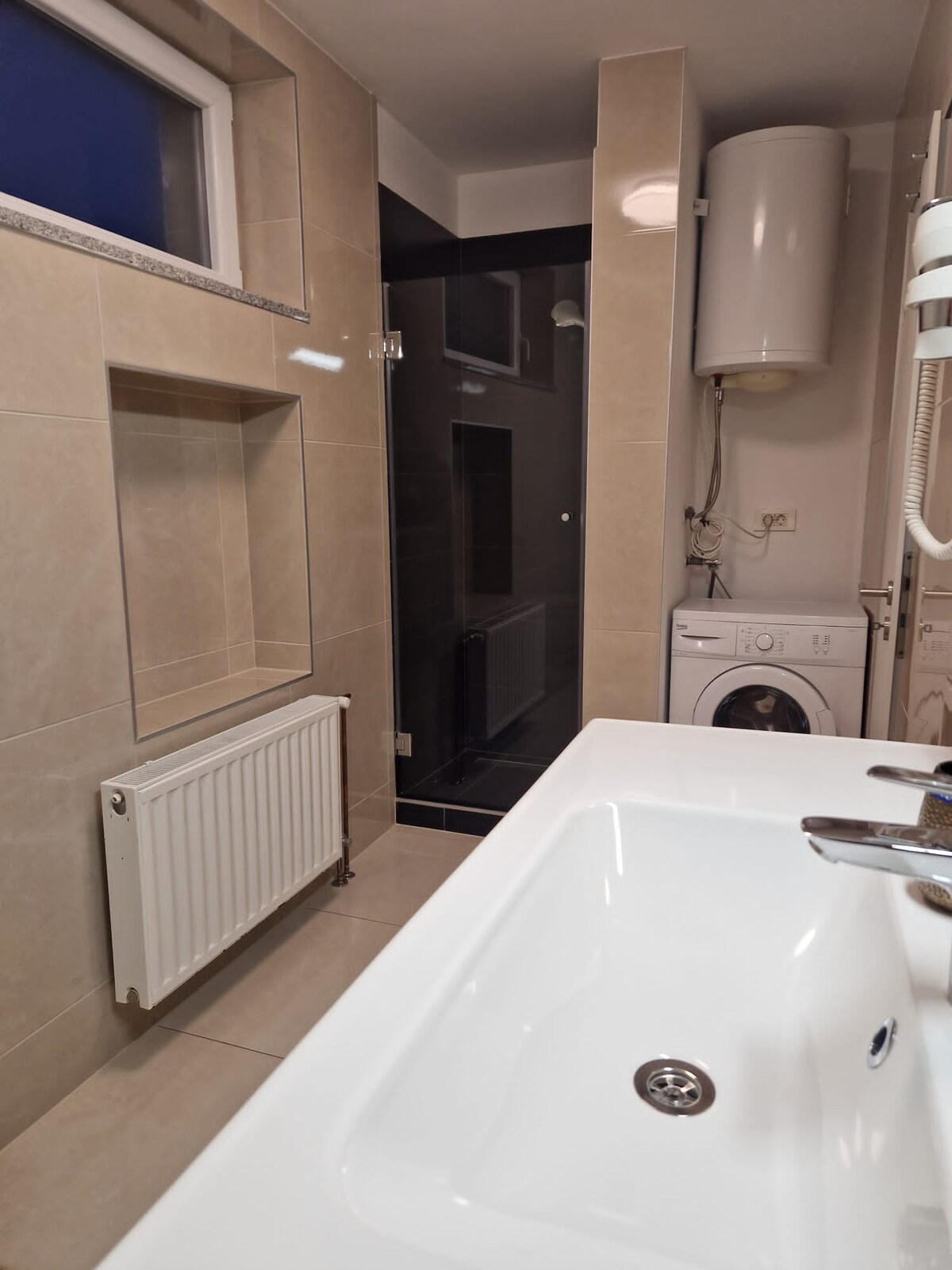 Narrow bathroom in cream tile with a recessed shelf, a radiator and a basin in the foreground, leading to a dark grey tiled shower, with a washing machine and water heater to the right