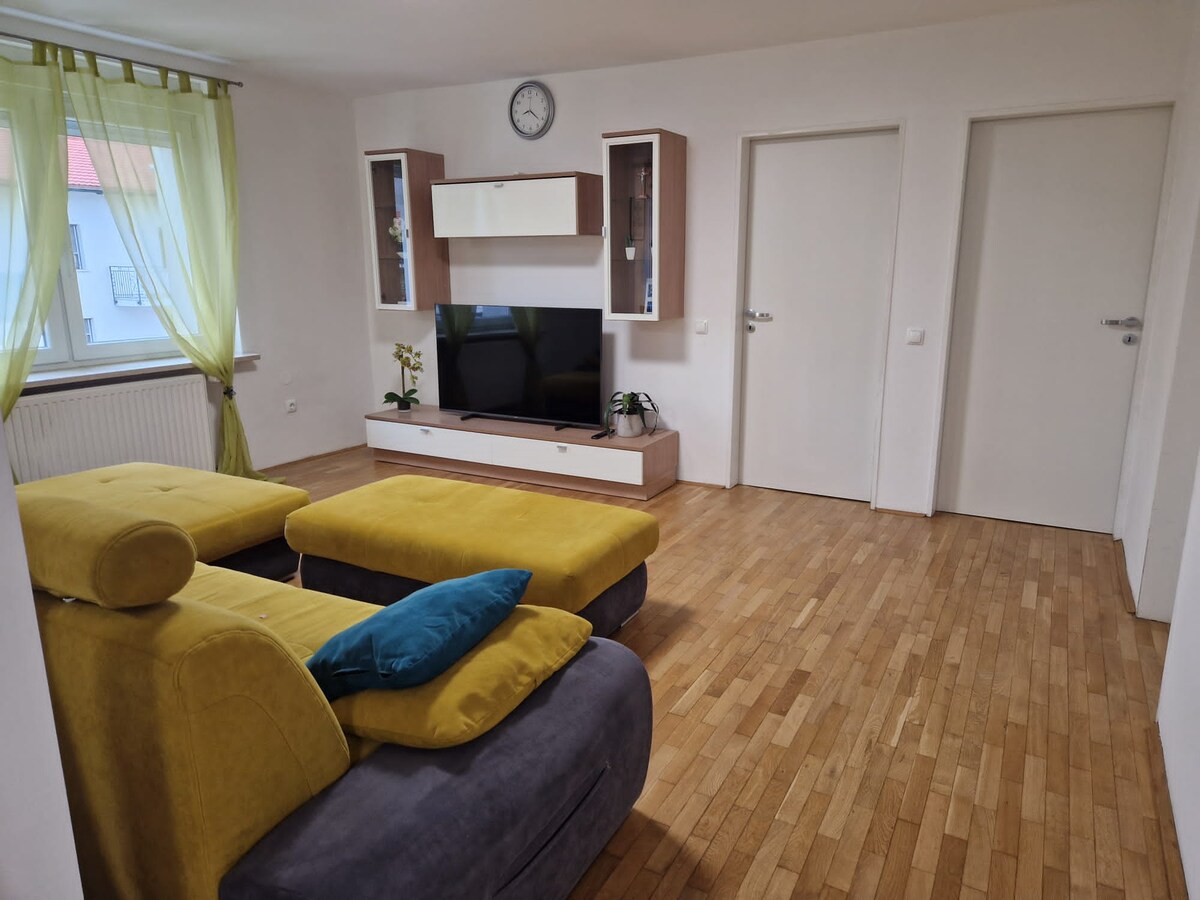 Living area with a flat-screen television on a white and oak sideboard, an orchid beside it, a wall clock above and two closed white doors on the right