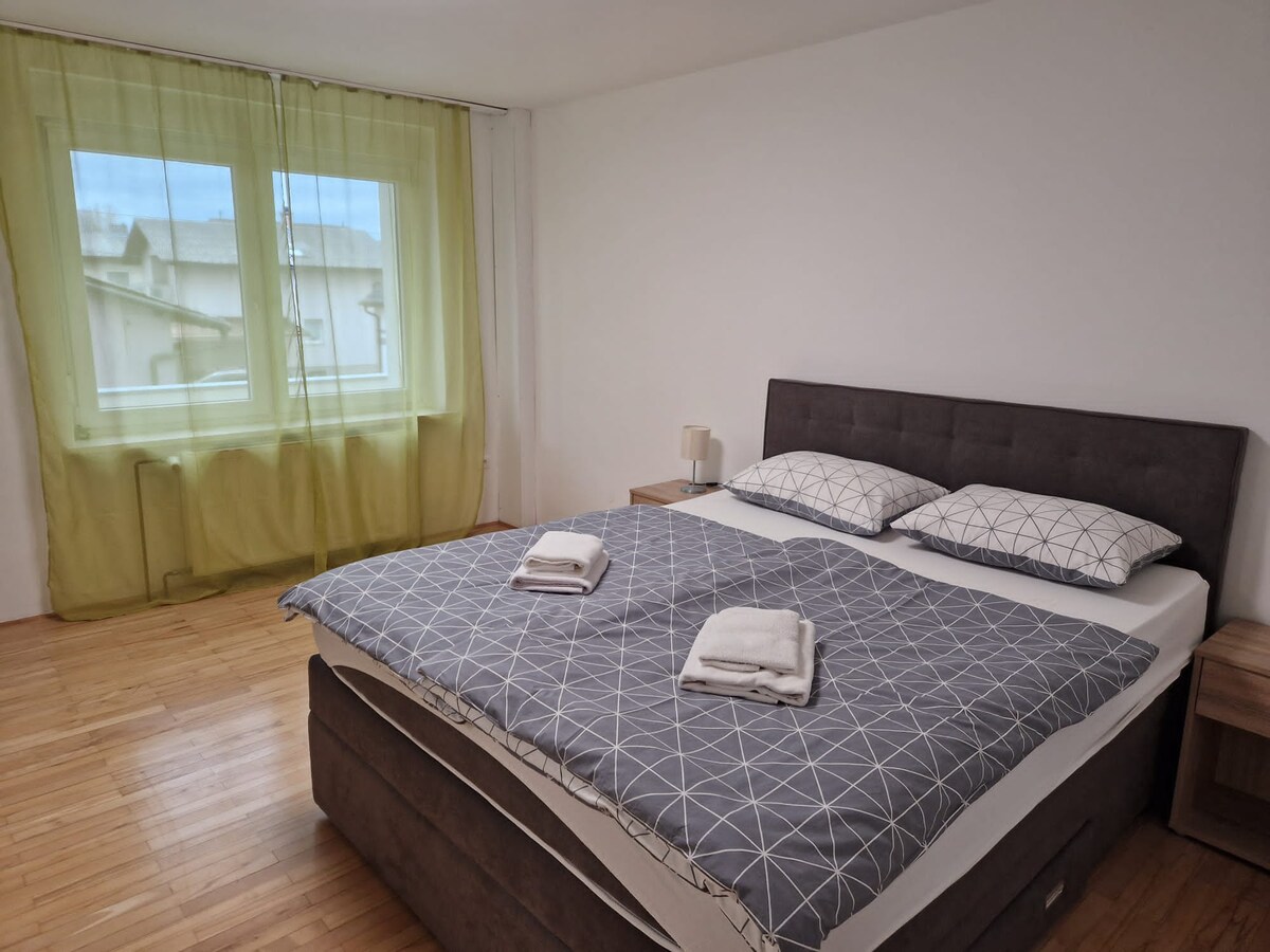 Bedroom with a grey upholstered double bed dressed in grey geometric bedding, bedside tables and a lamp either side, and lime green voile curtains at a large window