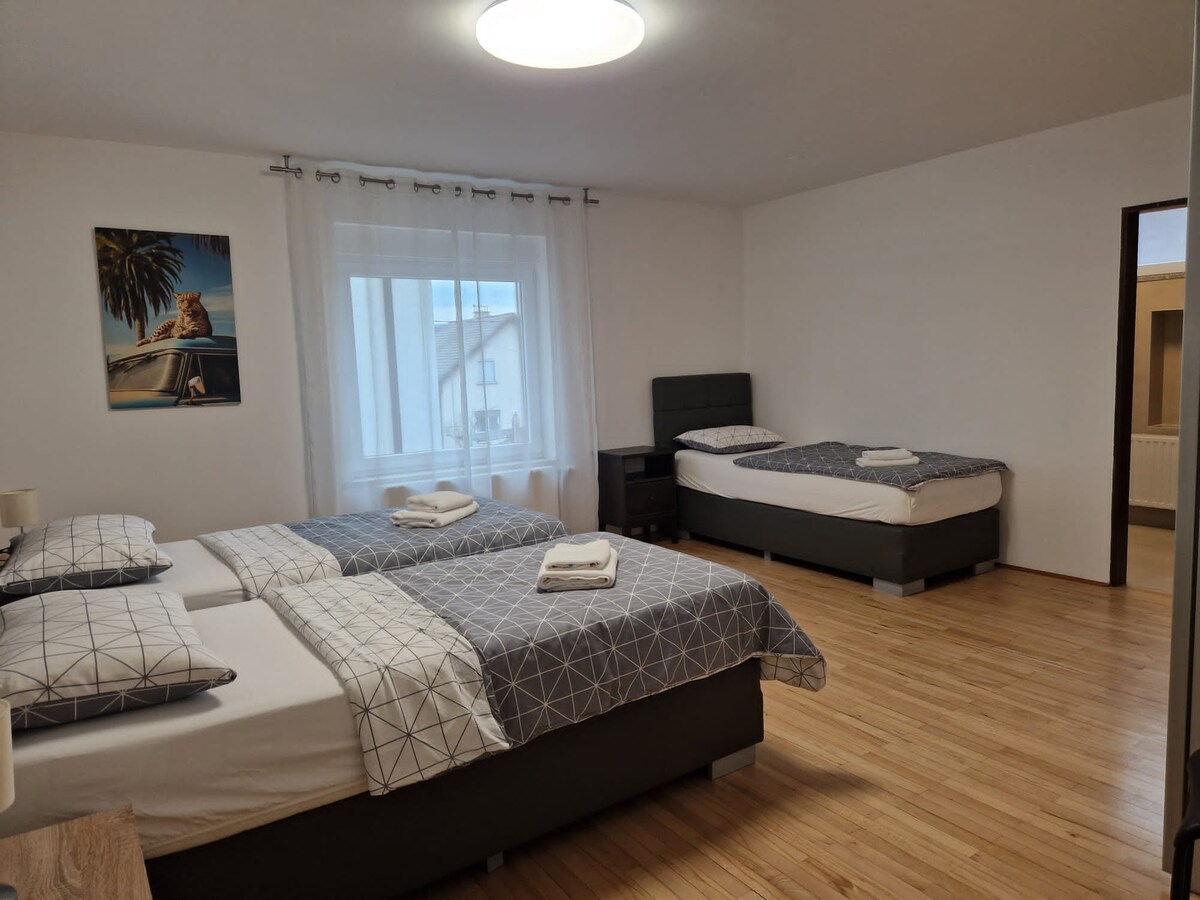 Spacious bedroom with a double bed dressed in grey geometric bedding in the foreground and a separate single bed with a dark headboard against the far wall, white curtains at the window and a leopard print canvas