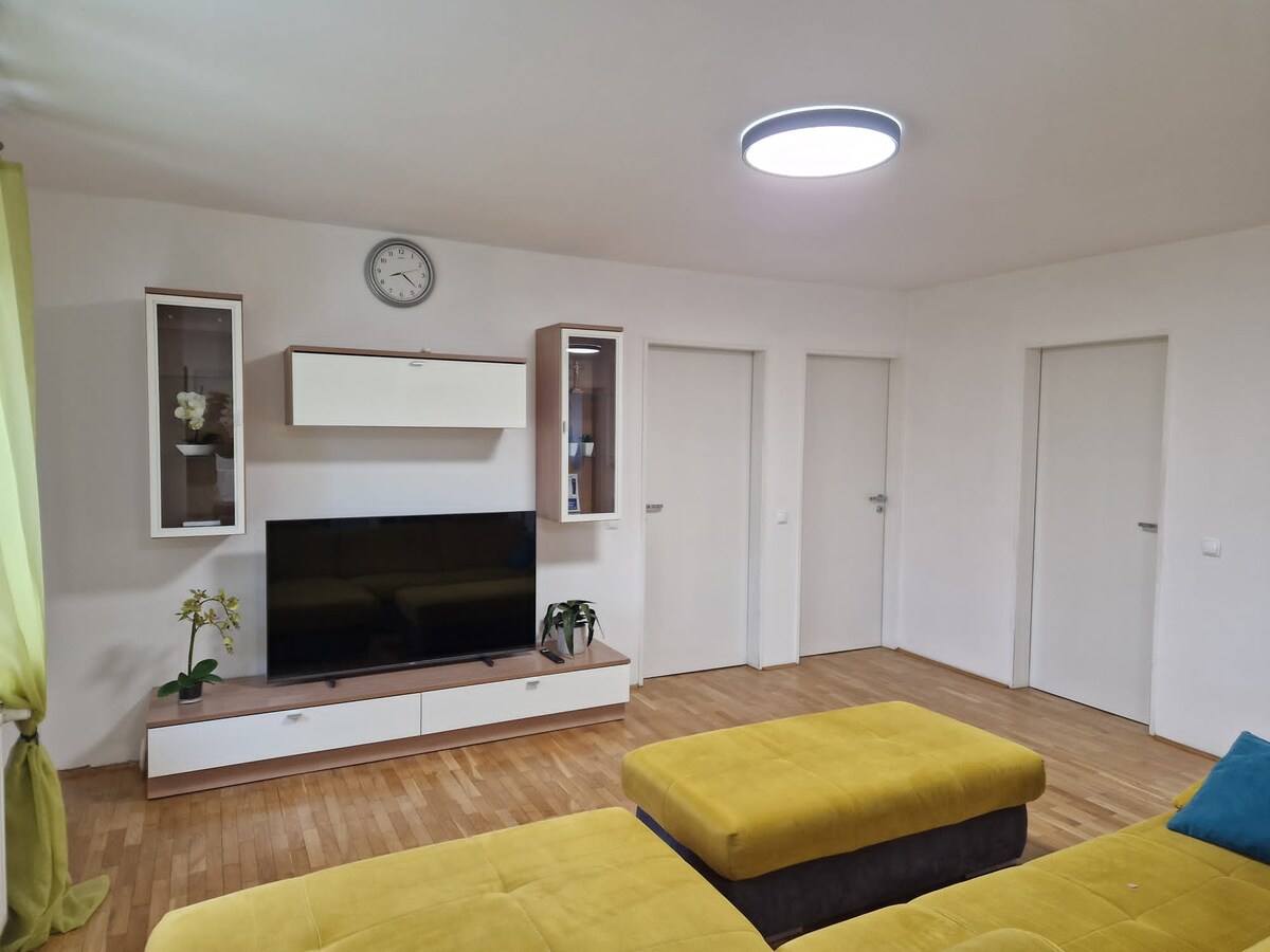 Living area with a wall-mounted television on a white and oak sideboard, two glass display cabinets and a clock above, three white doors along the right wall and a yellow sofa in the foreground