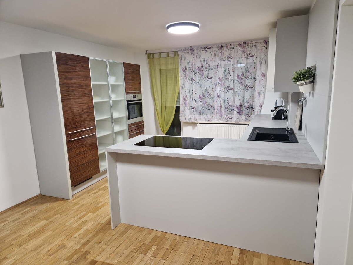 Kitchen seen from the living room: a light grey peninsula with an induction hob facing into the room, wall cupboards behind, a kettle and coffee machine on the worktop and a wooden floor