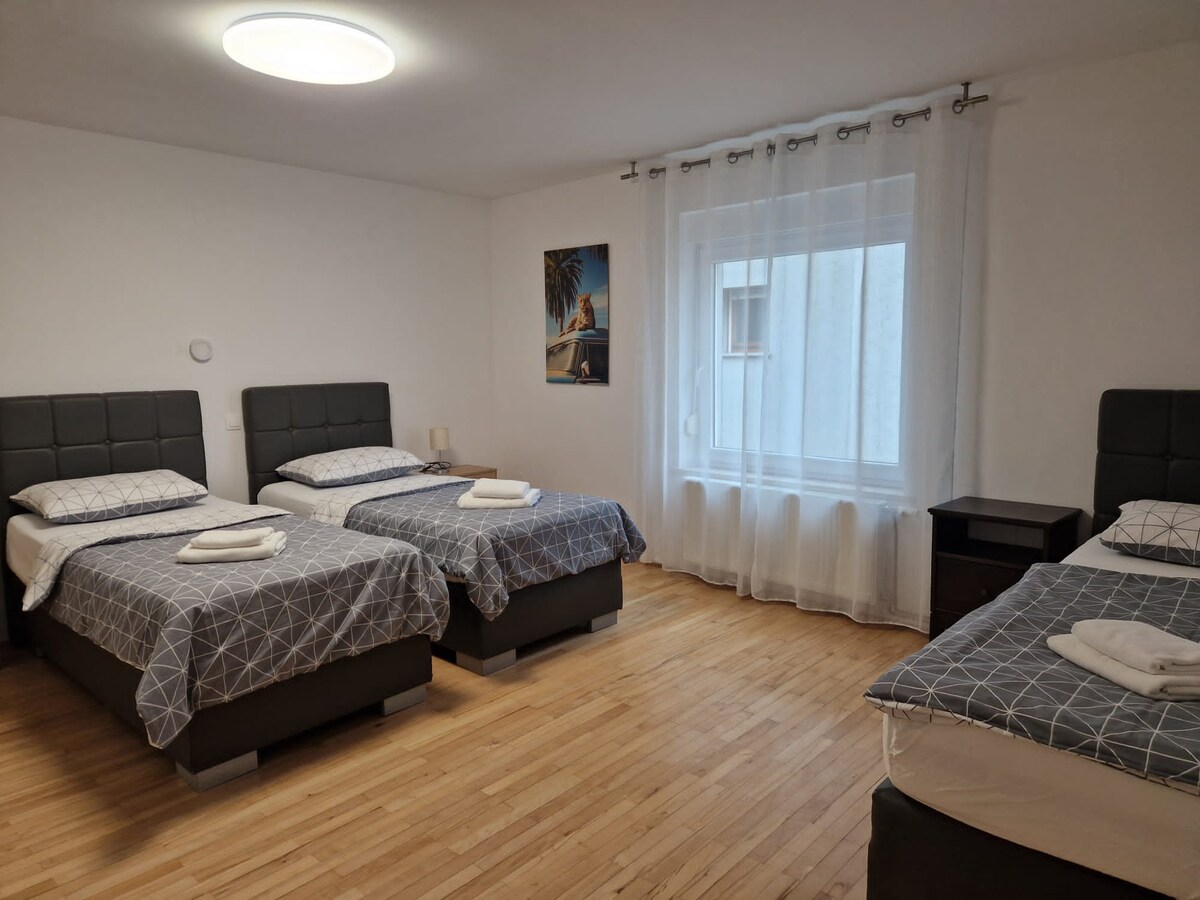 Large bedroom containing three single beds with dark padded headboards and matching grey geometric bedding, folded towels on each, a bedside table and a bright curtained window