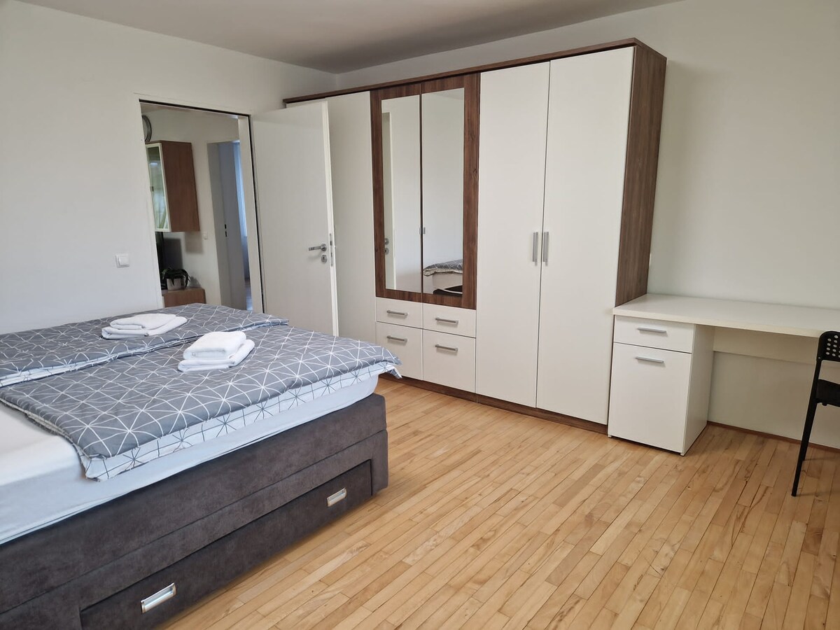 Bedroom with a grey upholstered double bed with storage drawers, a large white and walnut wardrobe with a mirrored door, and a white desk with a black chair against the right wall