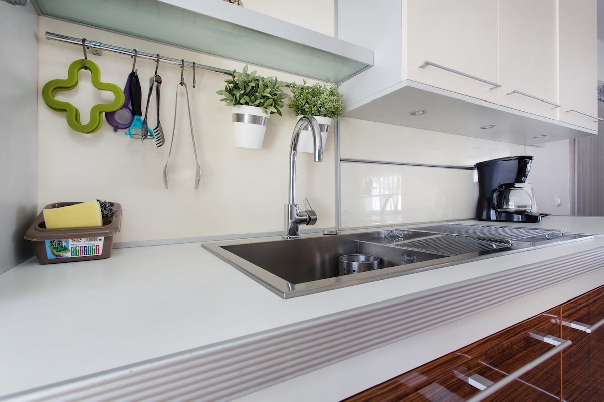 Close view of a stainless steel double sink and draining rack in a white worktop, with cooking utensils and potted herbs hanging from a rail and a filter coffee machine at the end
