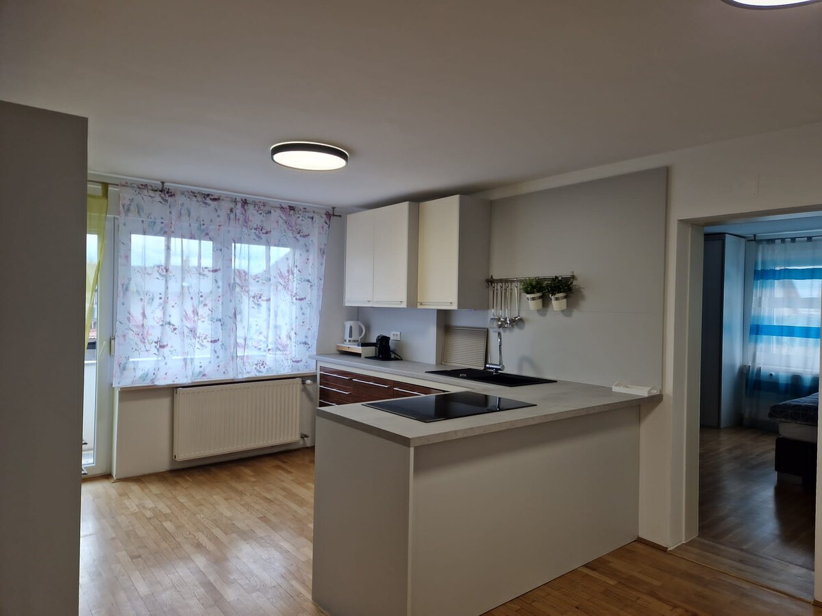 Kitchen with white wall cupboards, a light worktop with hob and black sink, floral curtains at the window, and a doorway on the right into a bedroom with blue striped curtains
