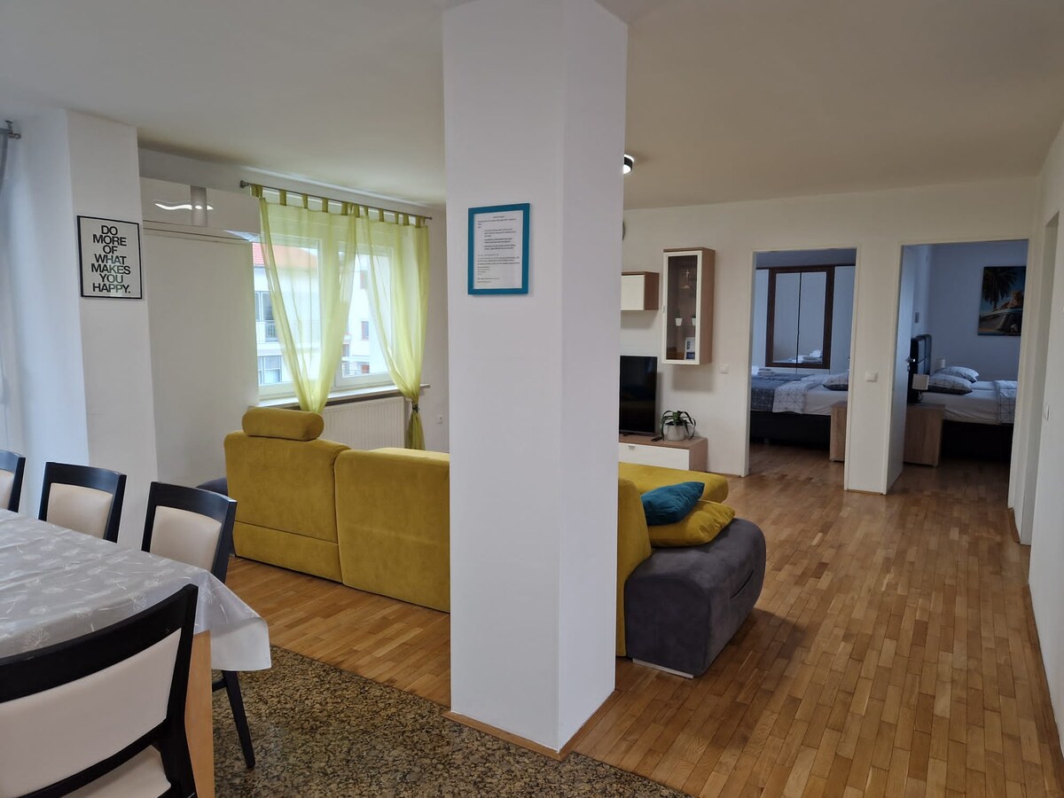 Long view through the apartment past dining chairs and the yellow sofa, with two bedrooms visible through open doorways at the far end, each containing beds
