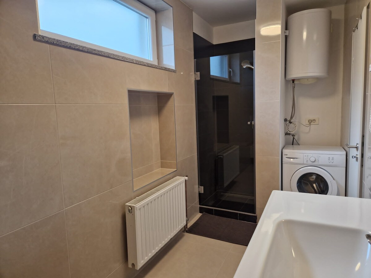 Cream tiled bathroom with a frosted window, a recessed niche and a radiator, opening into a dark grey tiled shower, with a washing machine and boiler in the alcove on the right