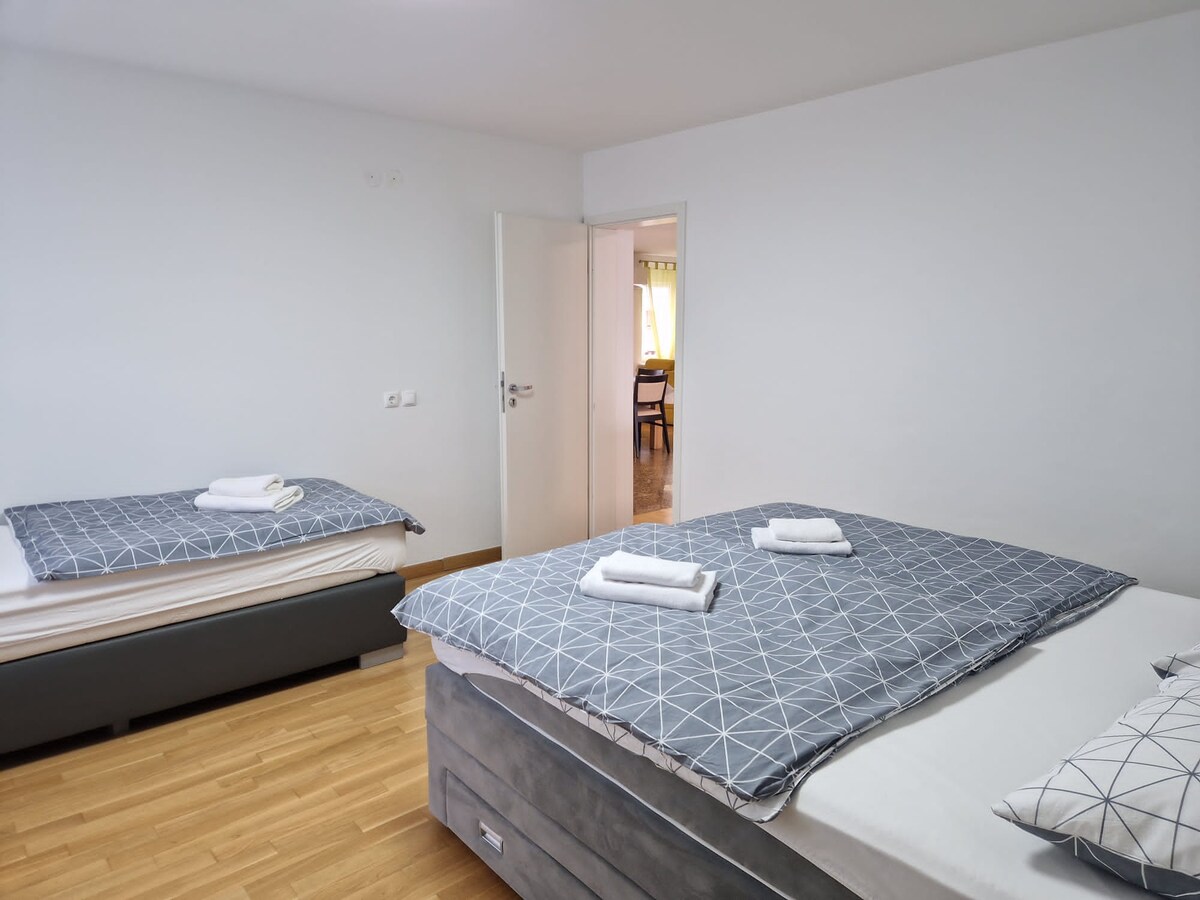 Plain white bedroom with a single bed on the left and a double bed on the right, both with grey geometric covers and folded towels, and an open door showing the dining area beyond