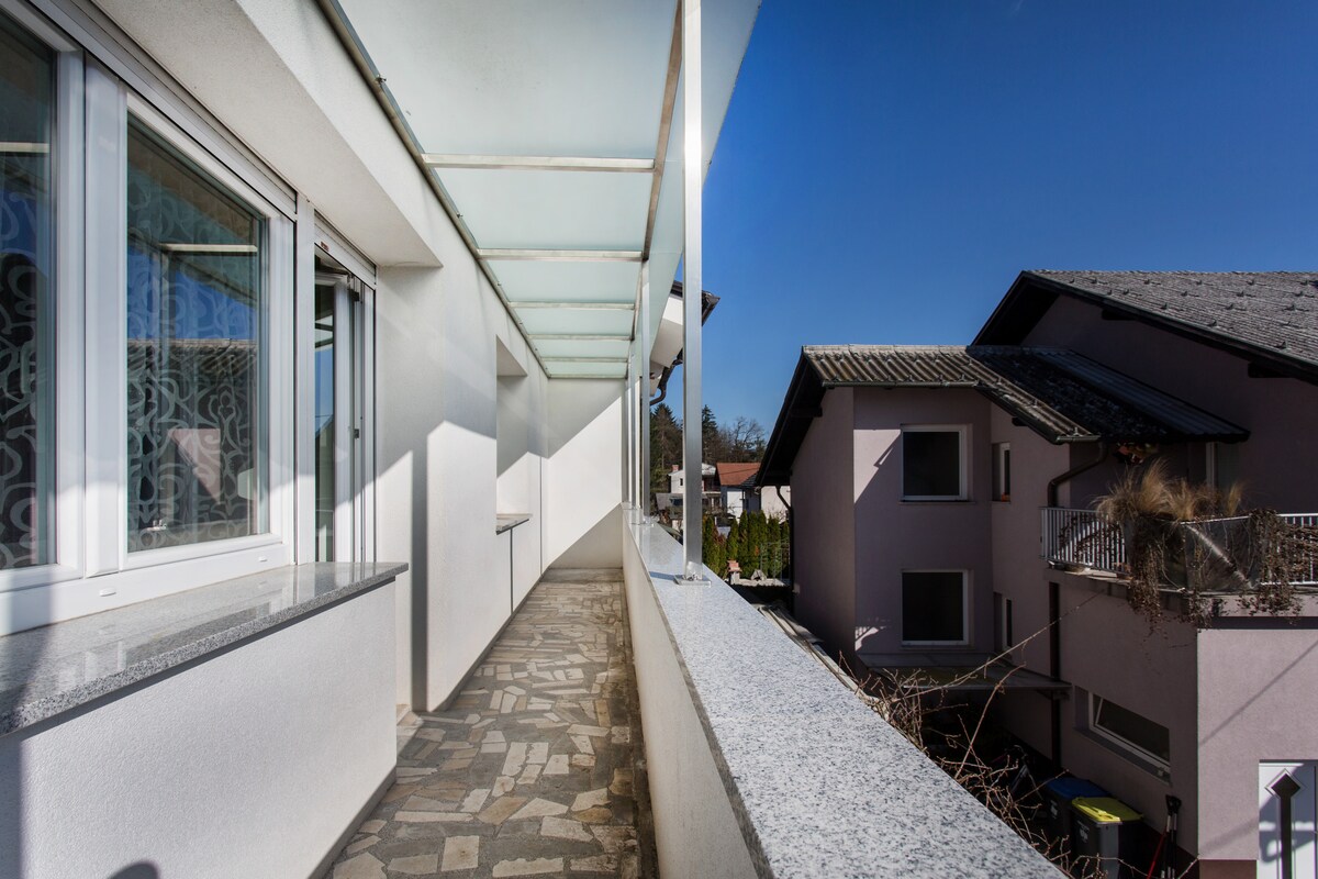 A long covered terrace with a flagstone floor and a granite handrail running past the apartment windows, looking out over a neighbouring house and rooftops under a clear blue sky