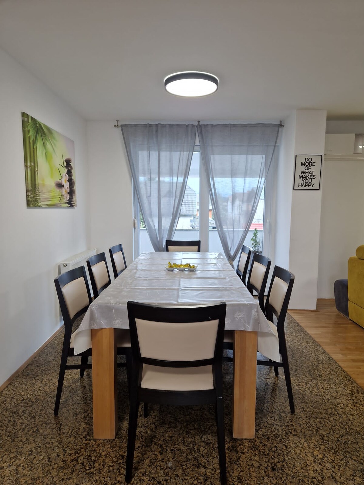 A long wooden dining table under a white cloth with eight black and cream chairs around it, a bowl on the table, tall grey curtains behind and a bamboo canvas on the left wall
