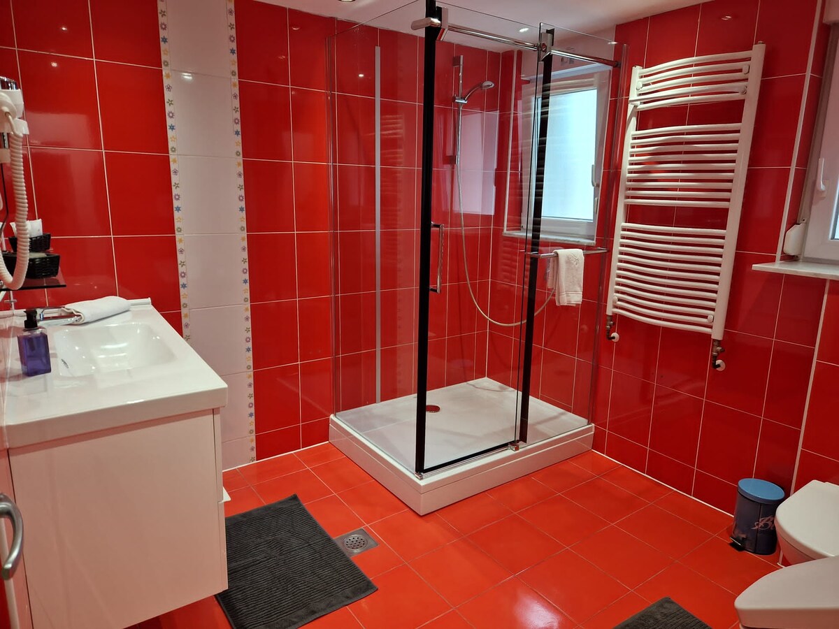 Bathroom tiled in bright red with a large glass shower cubicle, a white wall-hung vanity unit and basin, a white heated towel rail and a hairdryer on the wall