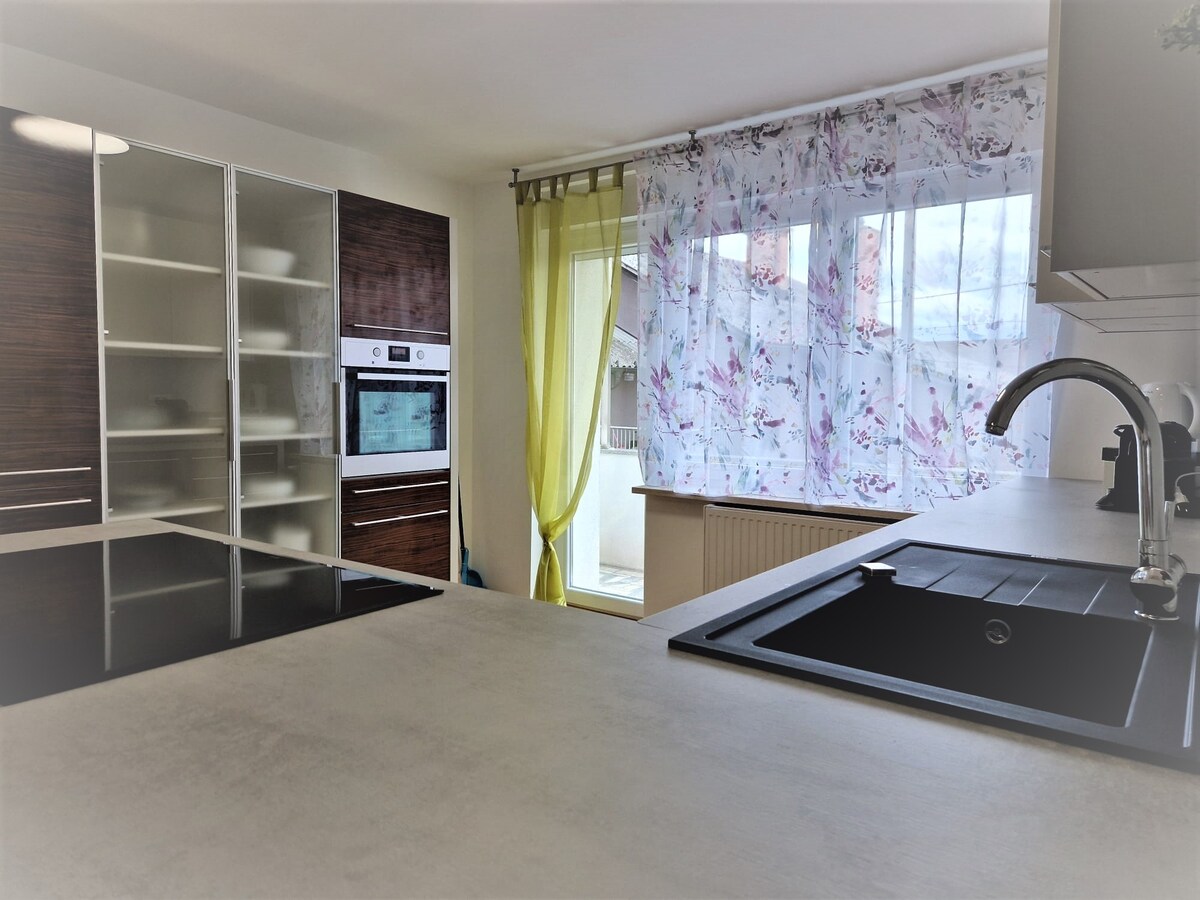 Kitchen corner with a built-in oven in a walnut tall unit, glass-fronted cupboards of crockery, a black sink and induction hob in a light worktop, and a glazed terrace door with green and floral curtains