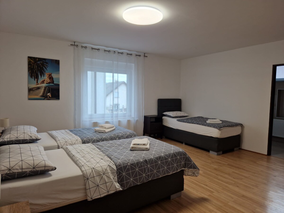 Bedroom with a double bed and a single bed on a wooden floor, both with grey patterned covers and folded towels, a leopard print canvas on the wall and a curtained window between them