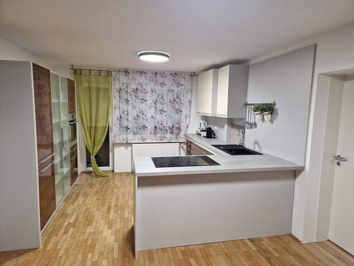 Wide view of an L-shaped kitchen with white wall cupboards, a concrete-look worktop with induction hob and black sink, herbs hanging from a rail and a terrace door with lime green curtains