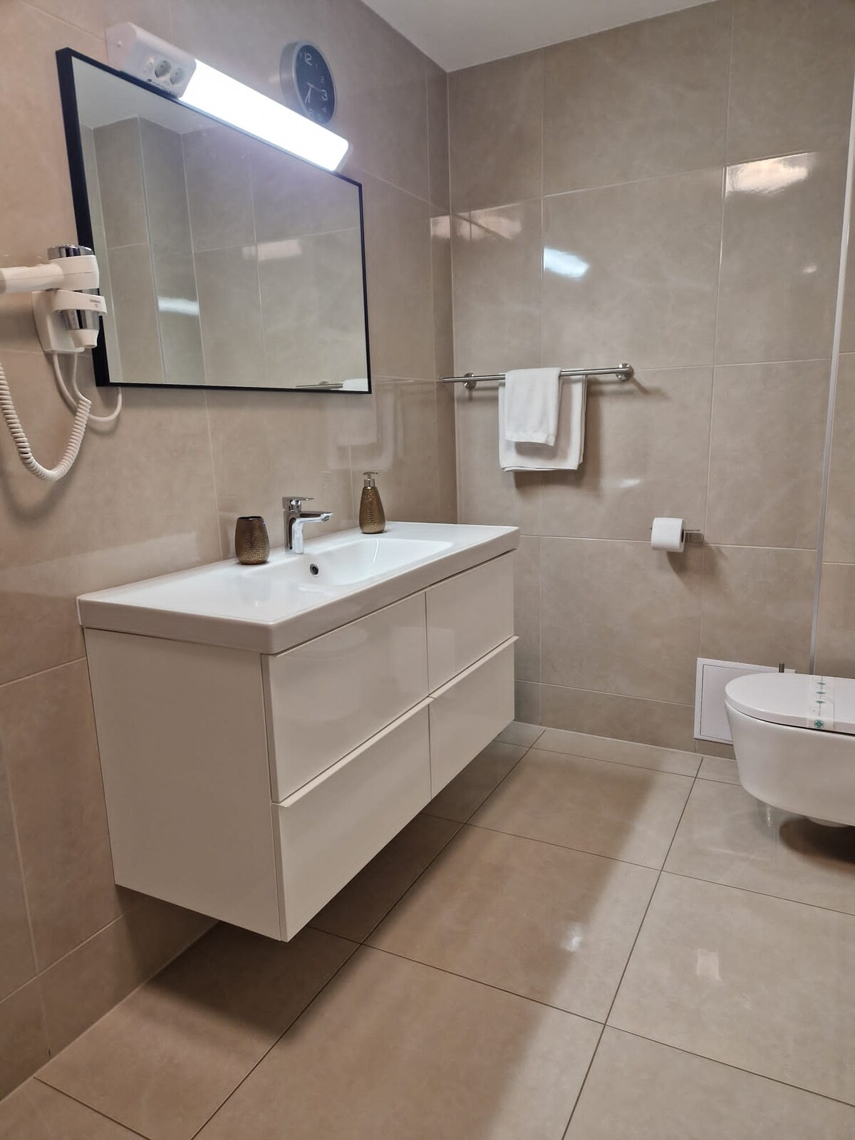 Cream stone-tiled bathroom with a wide white gloss vanity unit and basin under a black-framed mirror, a hairdryer on the wall, a towel rail and a wall-hung toilet