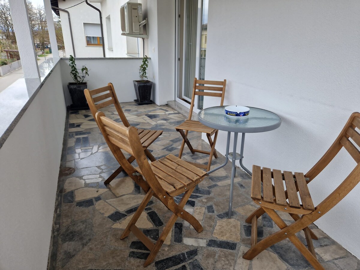 A round glass table with four folding wooden chairs on a flagstone terrace, potted plants along the granite parapet and an air-conditioning unit on the wall behind