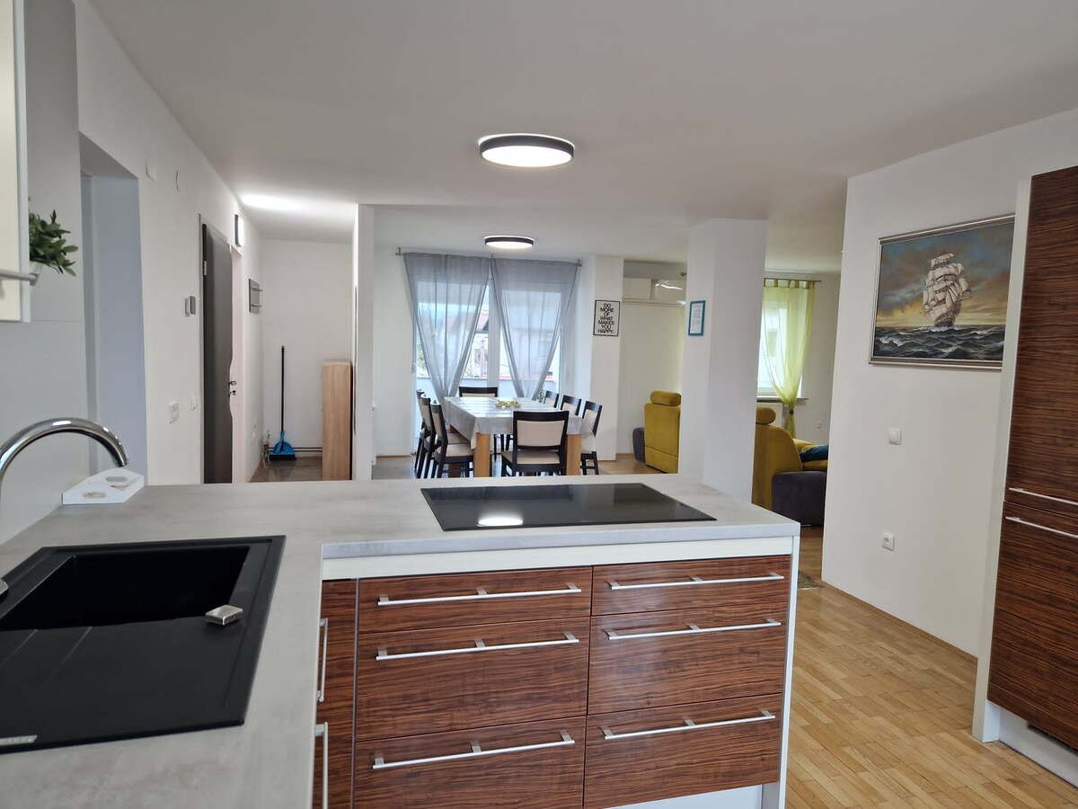 View from behind the kitchen peninsula with a black sink and induction hob set into a concrete-look worktop over walnut drawers, looking down the apartment to the dining table and yellow sofa