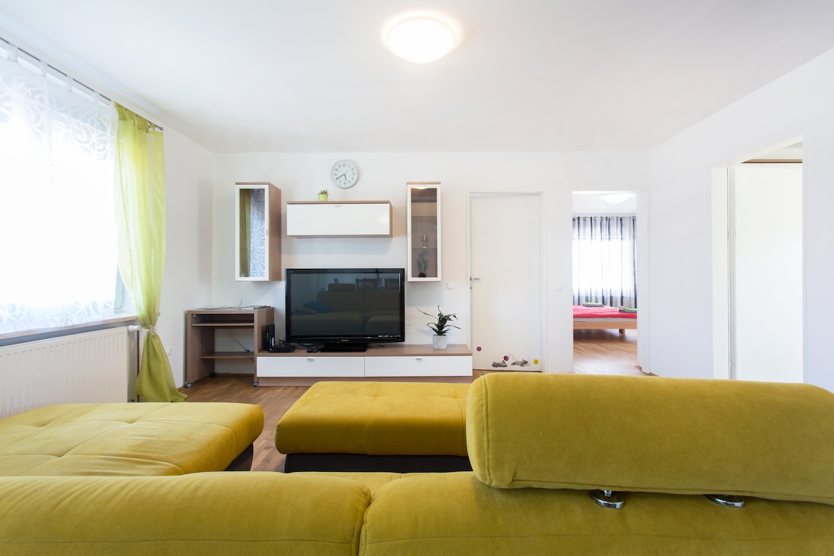 Living room with the yellow sofa in the foreground and a television opposite, with an open doorway on the right revealing a bedroom with a red bed