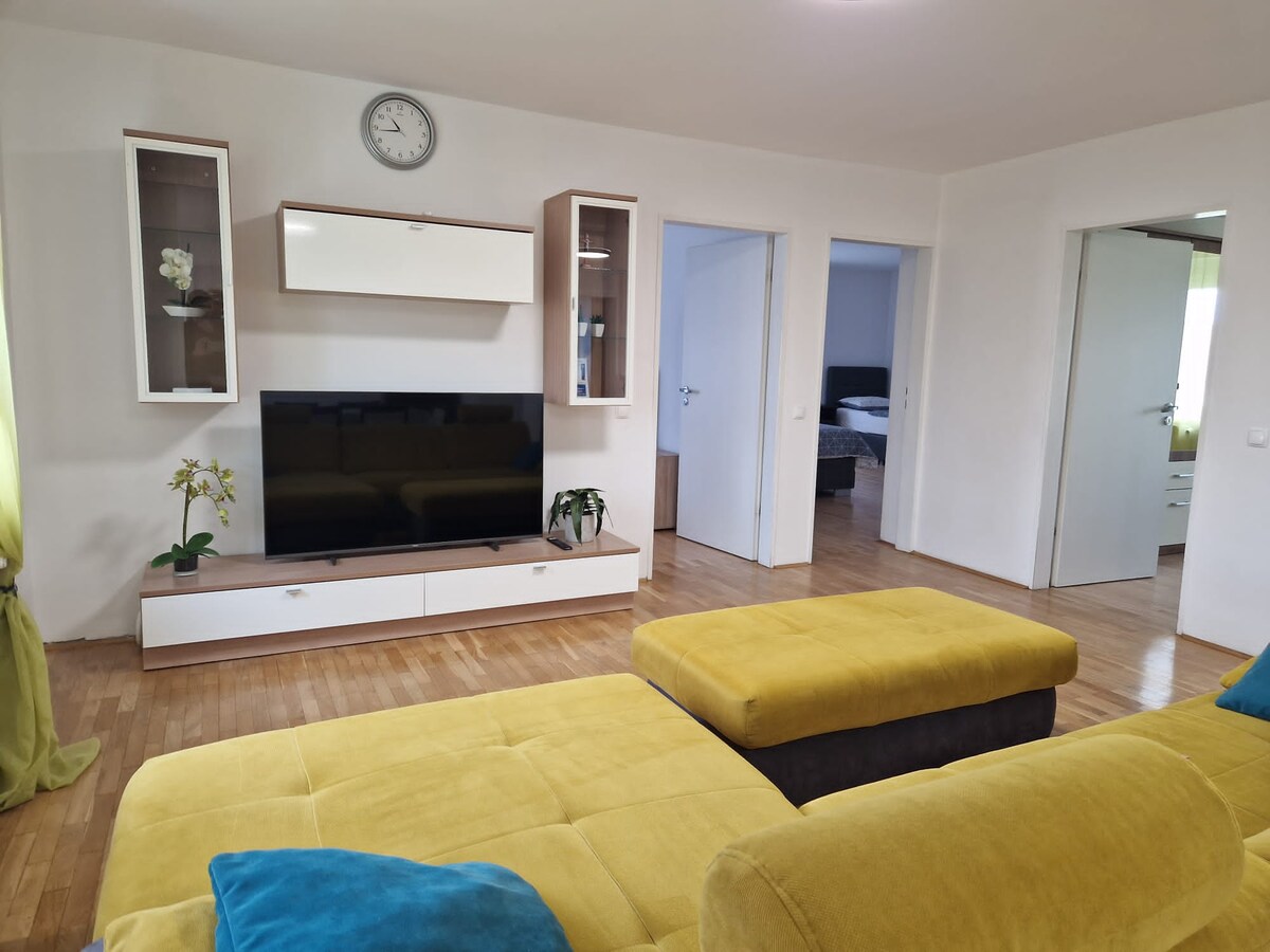 View across the yellow sofa to a television and shelving unit, with open doorways showing a bedroom with a grey bed on one side and a further room on the other