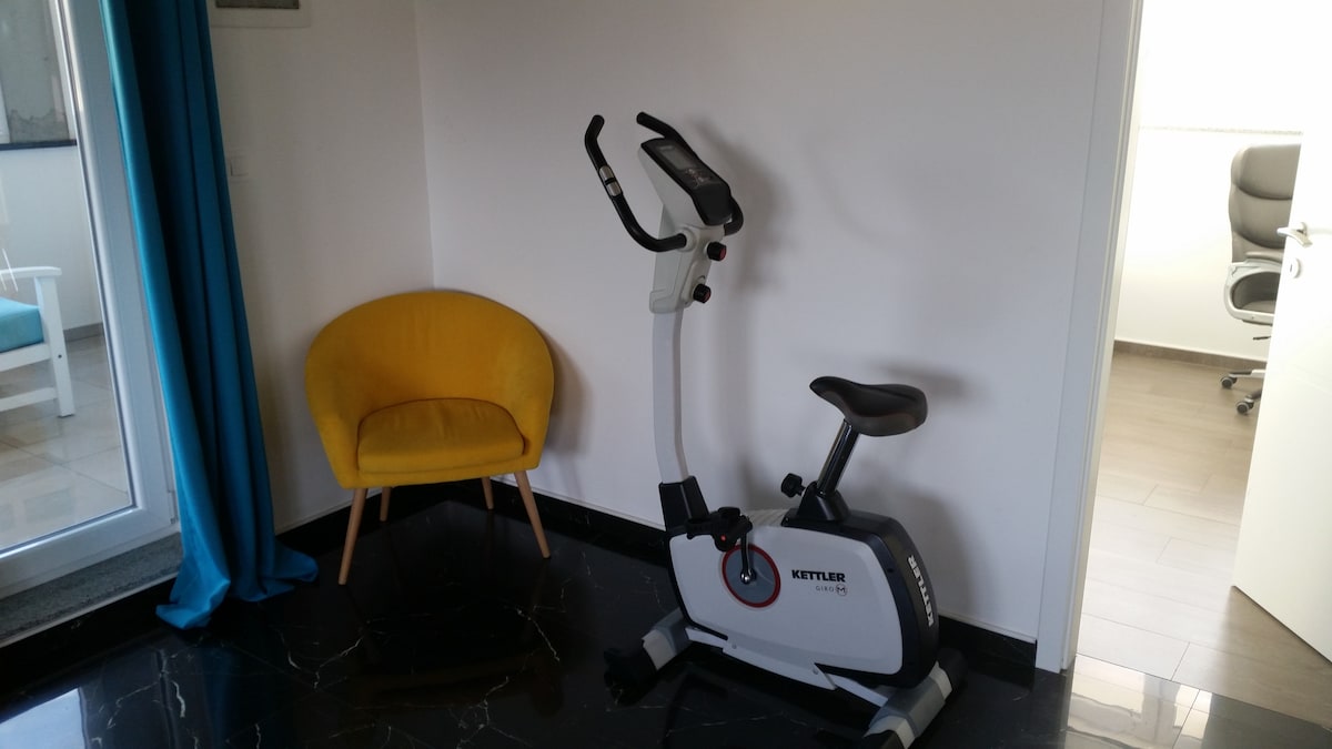 A yellow tub armchair beside an upright exercise bike against a white wall, on black marble, with a turquoise curtain at a glazed door on the left