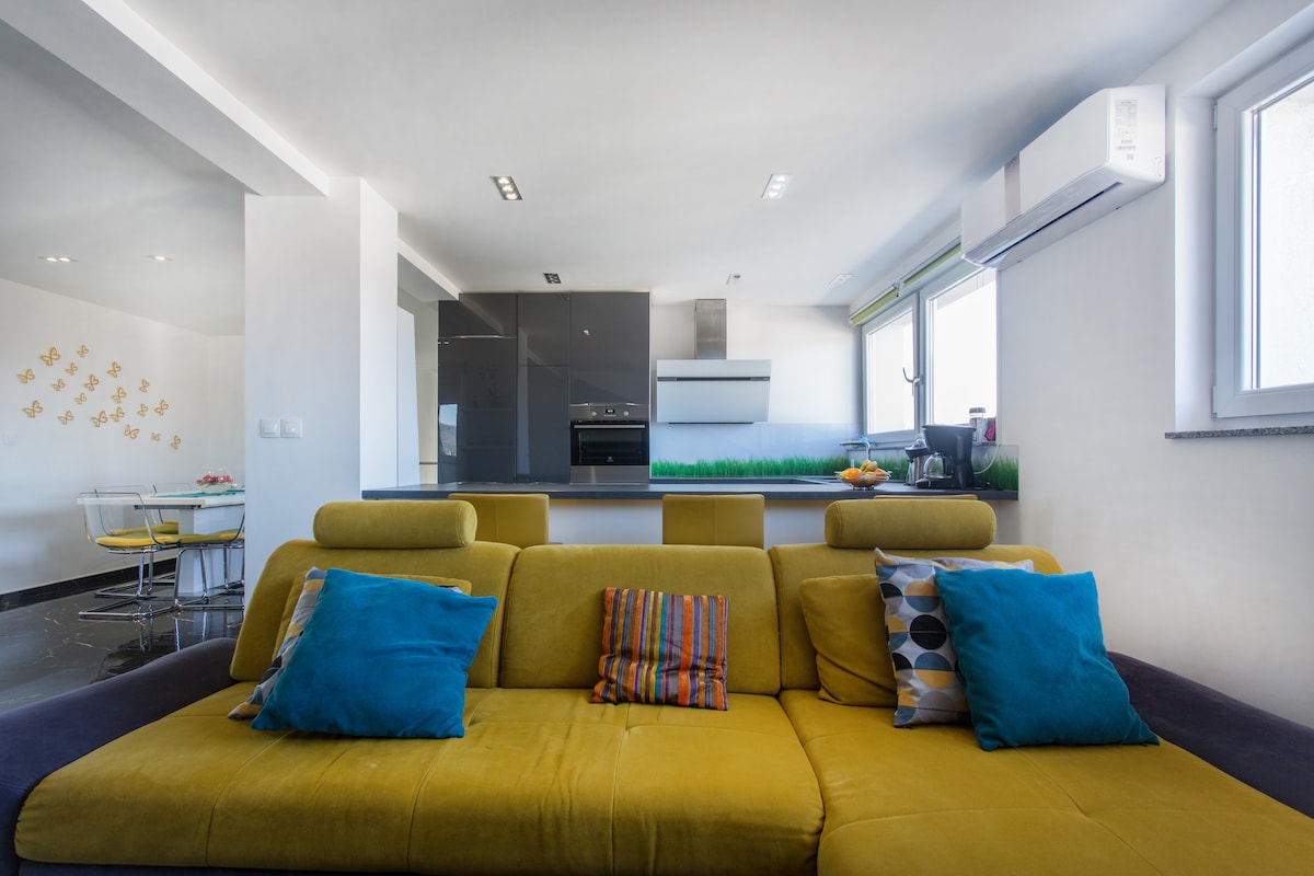 A long mustard sofa with teal and striped cushions facing the kitchen across a dark breakfast bar, with a white angled extractor hood and a green grass-print glass splashback, and an air-conditioning unit on the wall above