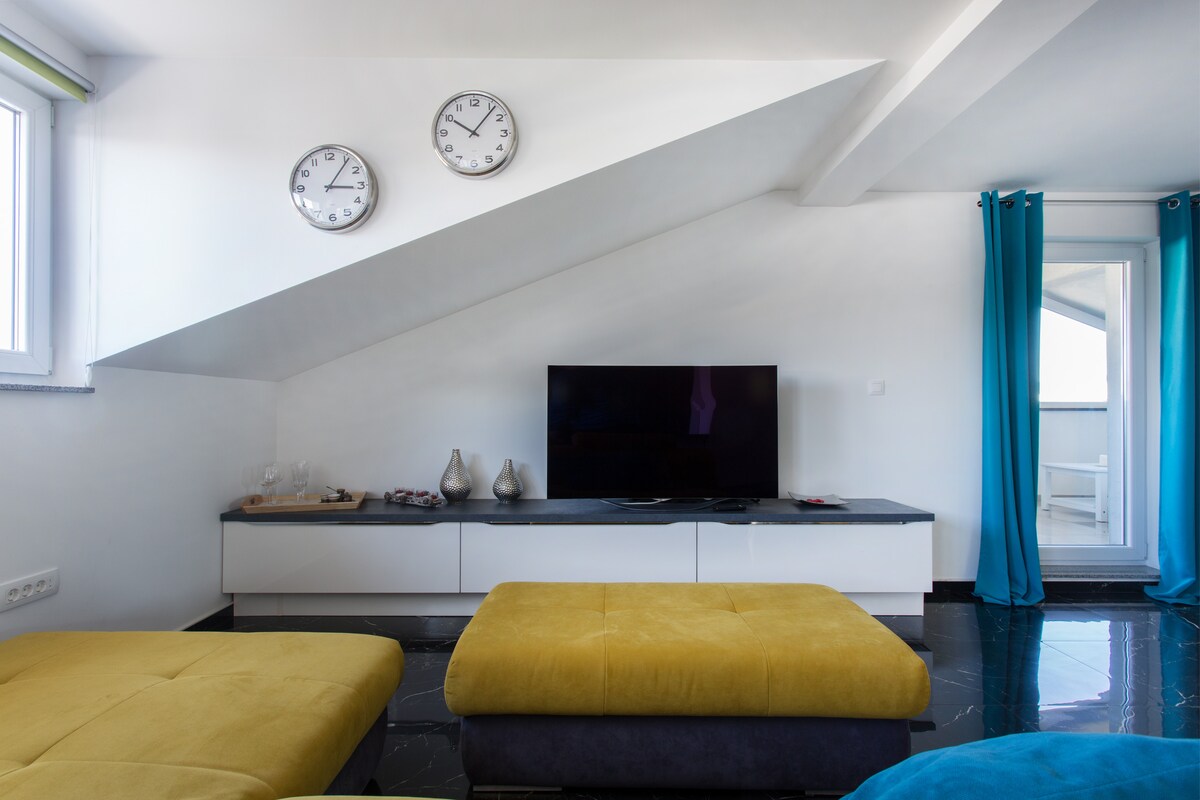 Living room with a steeply sloping white ceiling above a long low sideboard and television, two steel clocks on the wall, mustard seating in the foreground and full-length turquoise curtains at a terrace door