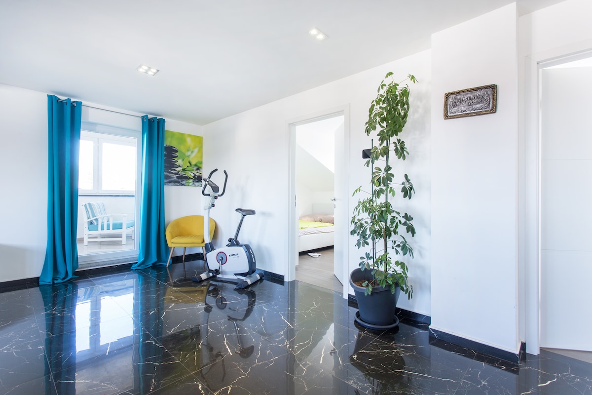 Bright top-floor room with a mirror-like black marble floor, turquoise curtains at a terrace door, a yellow armchair and exercise bike, a large potted plant and an open door showing a bed in the next room