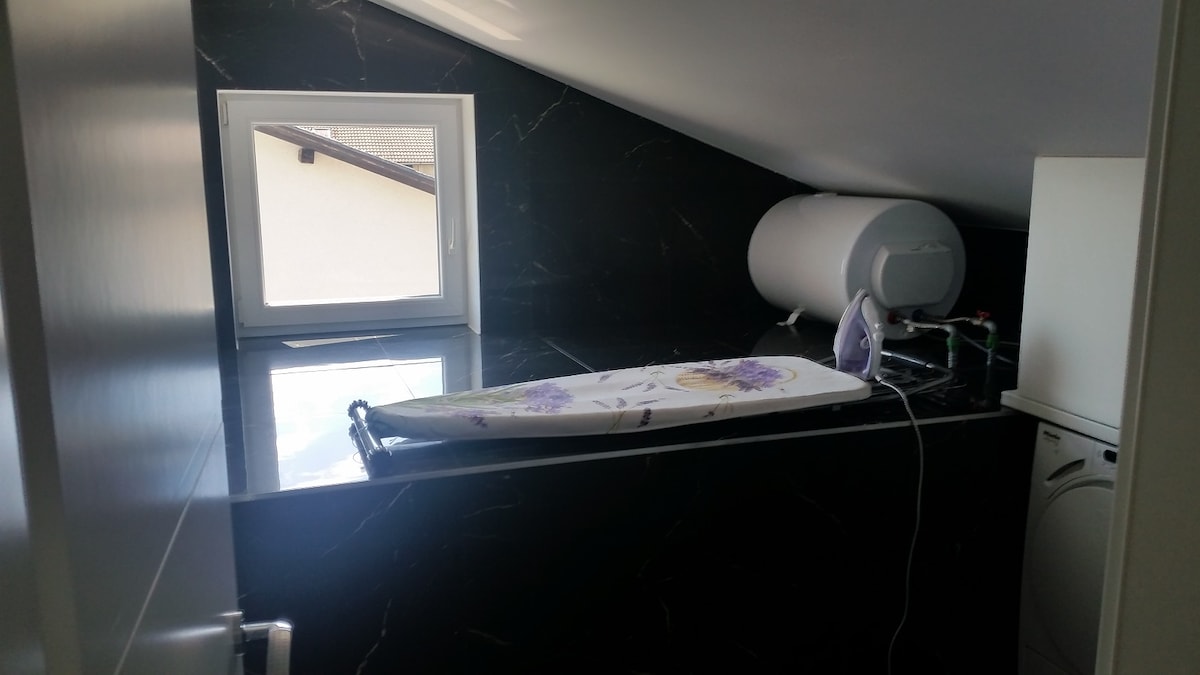 Utility space under the roof slope, tiled in dark marble, with an ironing board and iron set up, a wall-mounted water heater, a small window and a washing machine to the right