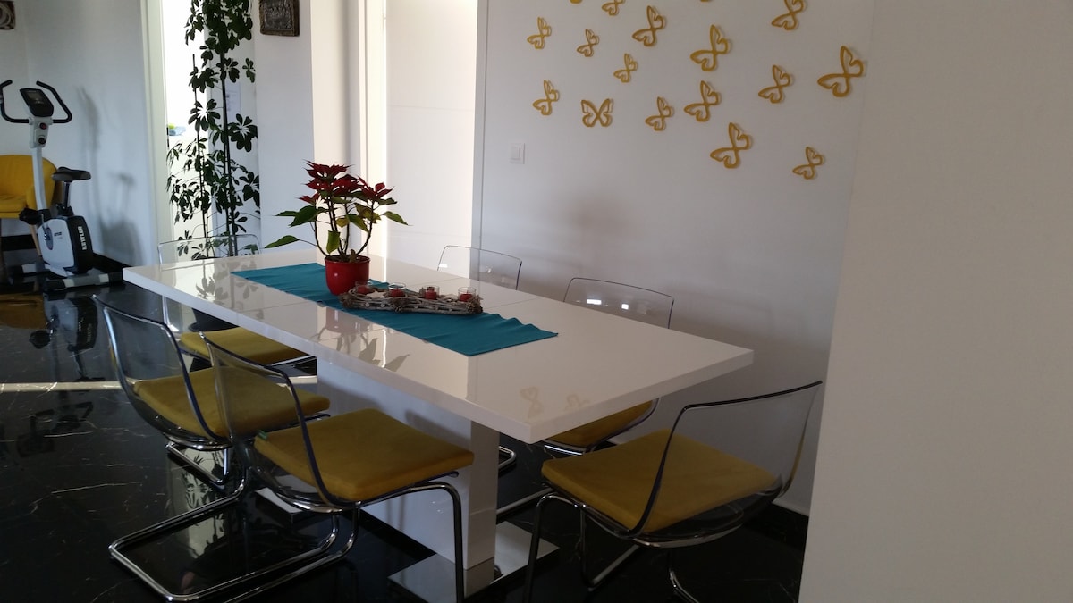 White gloss dining table set with a turquoise runner, a red poinsettia and candles, surrounded by transparent chairs with yellow cushions, with an exercise bike and a tall plant in the background