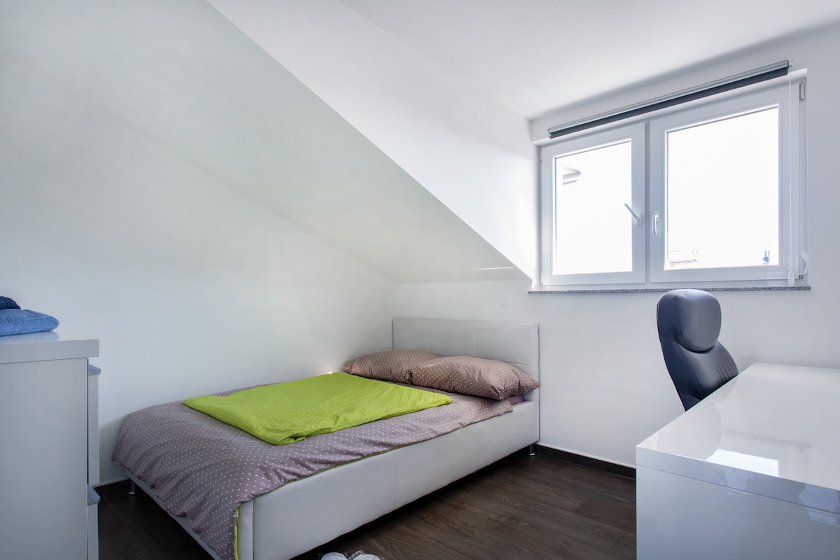 Small attic bedroom with a sloping white ceiling, a white bed with a lime green throw and spotted linen, a window with a roller blind, and a white gloss desk with a black office chair