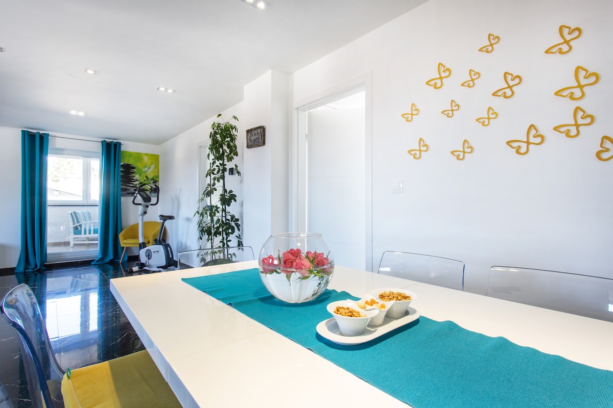 Dining table with a glass bowl of pink roses and bowls of nuts, gold butterflies on the wall to the right, and a turquoise-curtained door leading out to a chair on the terrace at the far end