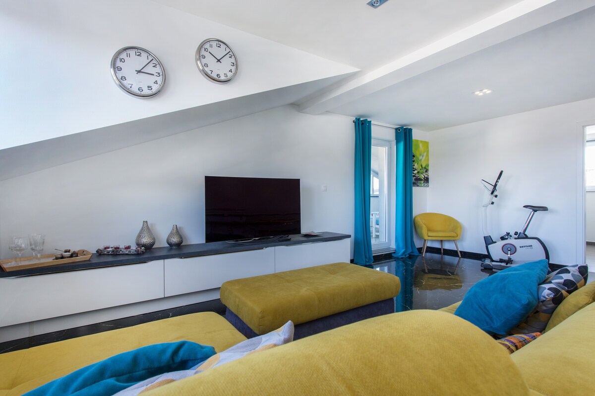 Living area with two round steel wall clocks, a large flat-screen television on a long white and dark stone sideboard, a mustard sofa and ottoman, and a turquoise-curtained terrace door beside an exercise bike