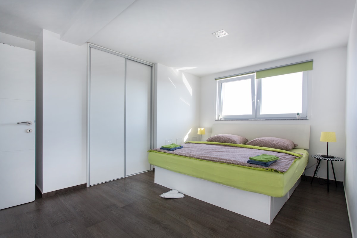 Bedroom seen from the doorway with a white double bed under lime green and mauve covers, bedside lamps, a window with a green blind and a tall white sliding wardrobe on the left, on dark wood flooring