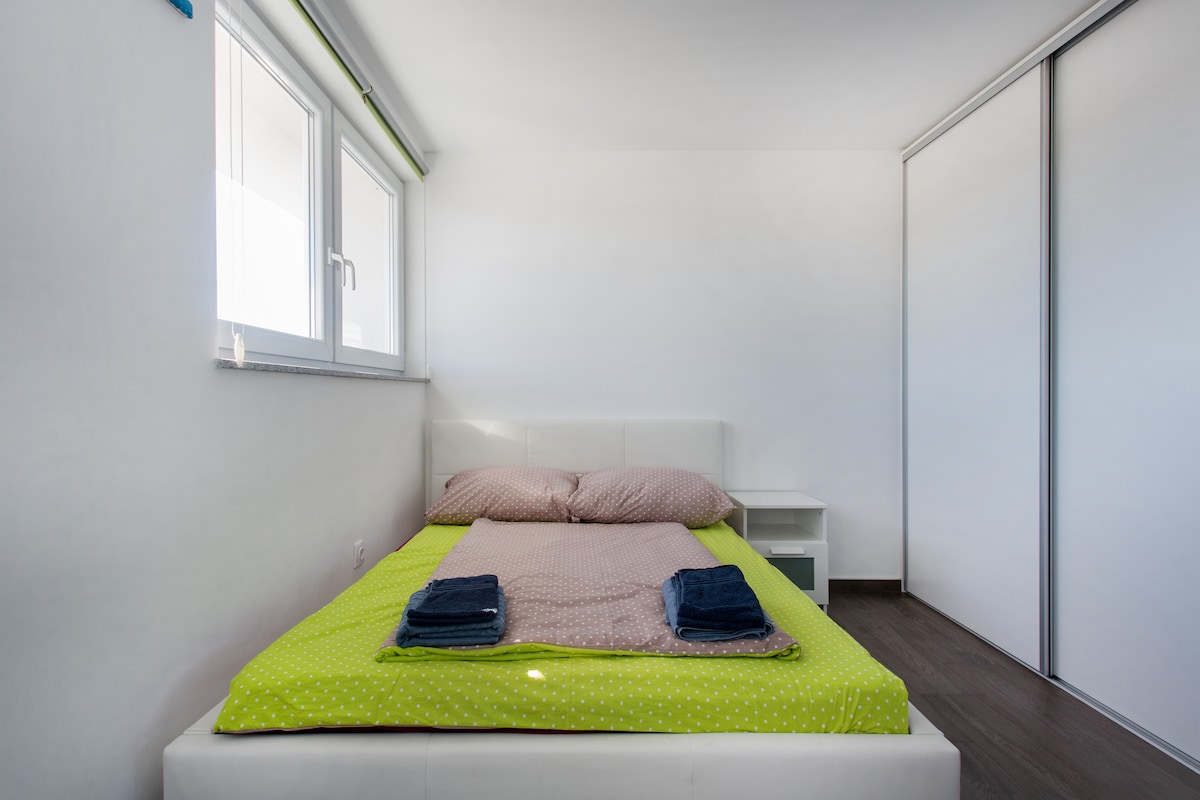 Bedroom with a white upholstered double bed under a lime green cover, folded blue towels on top, a small white bedside cabinet and a frosted glass sliding wardrobe filling the right-hand wall