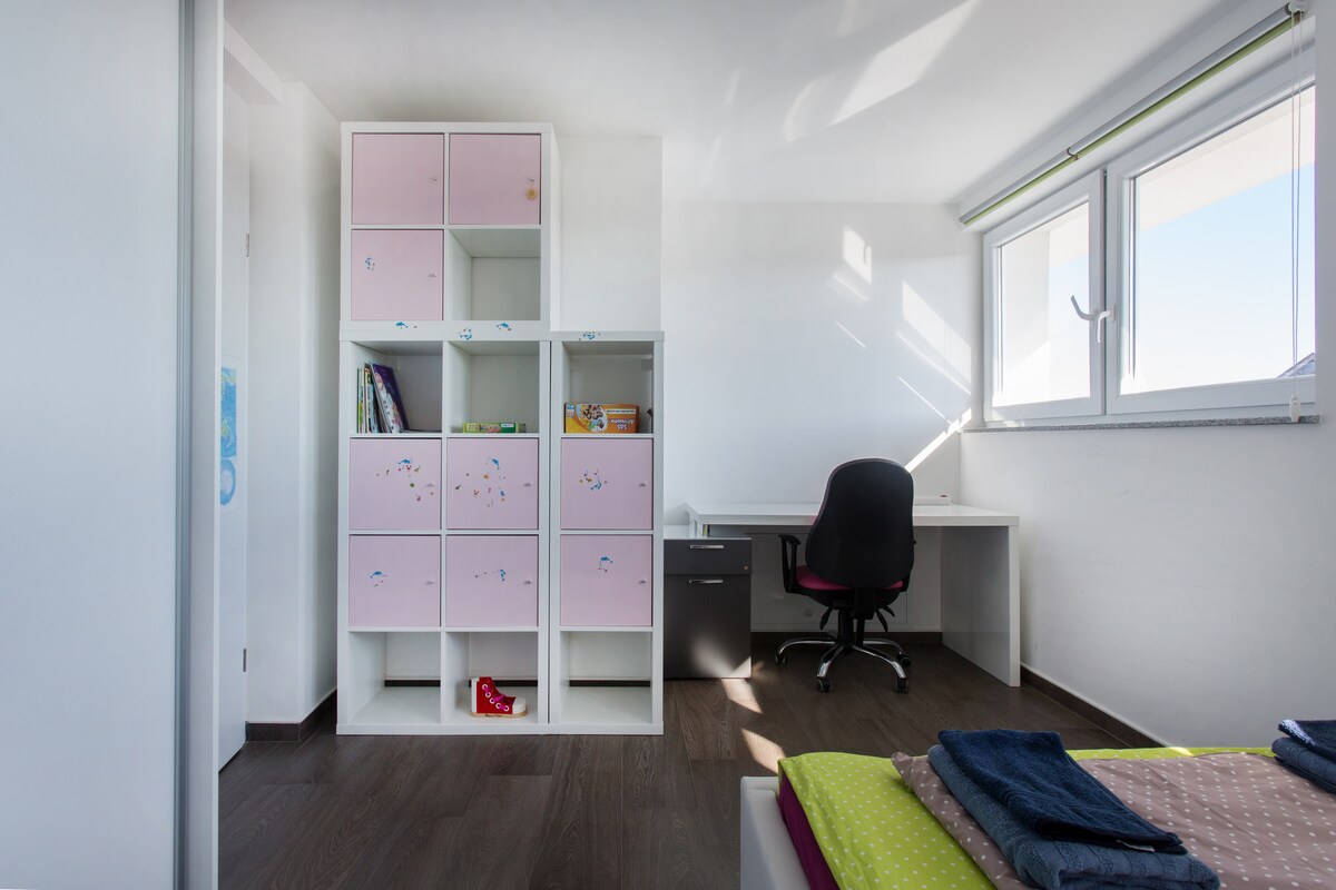 Child's bedroom with a white cube shelving unit holding pink fabric storage boxes, picture books and games, a white desk with a black chair beneath a bright window, and the corner of a bed in the foreground