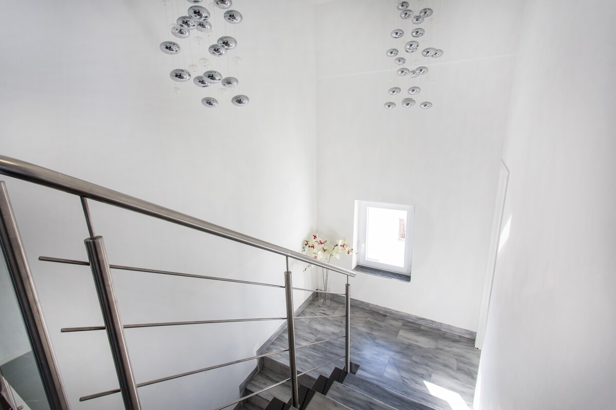 Looking down a stairwell with a brushed steel handrail and grey marble treads, white walls, a small window with a vase of orchids on the sill, and two clusters of mirrored glass ball pendant lights hanging from the high ceiling