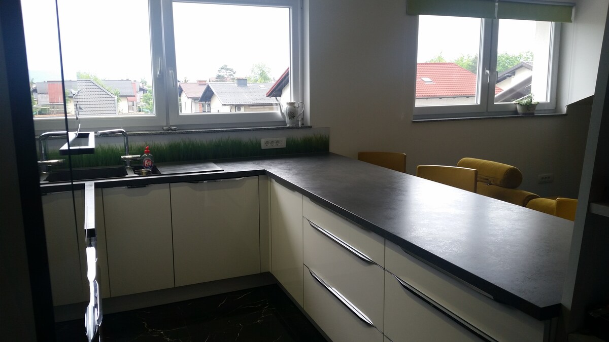 Kitchen worktop in dark laminate running under two windows that look out over tiled roofs and trees, with a stainless sink, cream drawer units and yellow bar stools at the far end