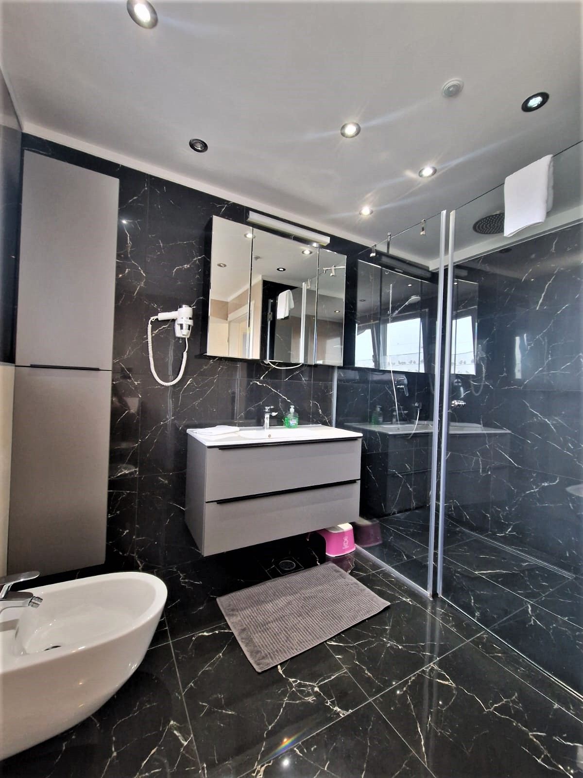 Bathroom lined in black marble with a wide white basin on a grey vanity unit, a mirrored cabinet above with a hairdryer beside it, and a glass-walled walk-in shower with a large round rain head