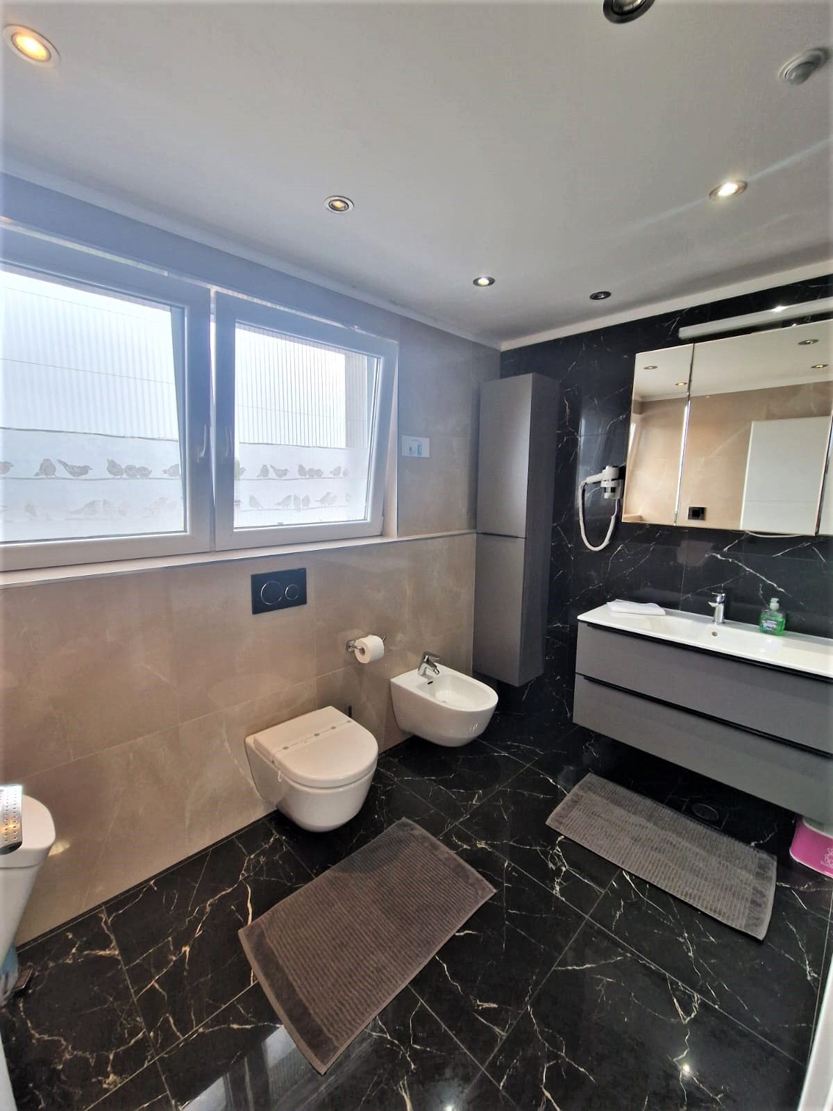 Wide view of the black marble bathroom with a grey tall cabinet and vanity unit, a white basin, a wall-hung toilet and bidet under the window, and a pink child's step stool beside the vanity