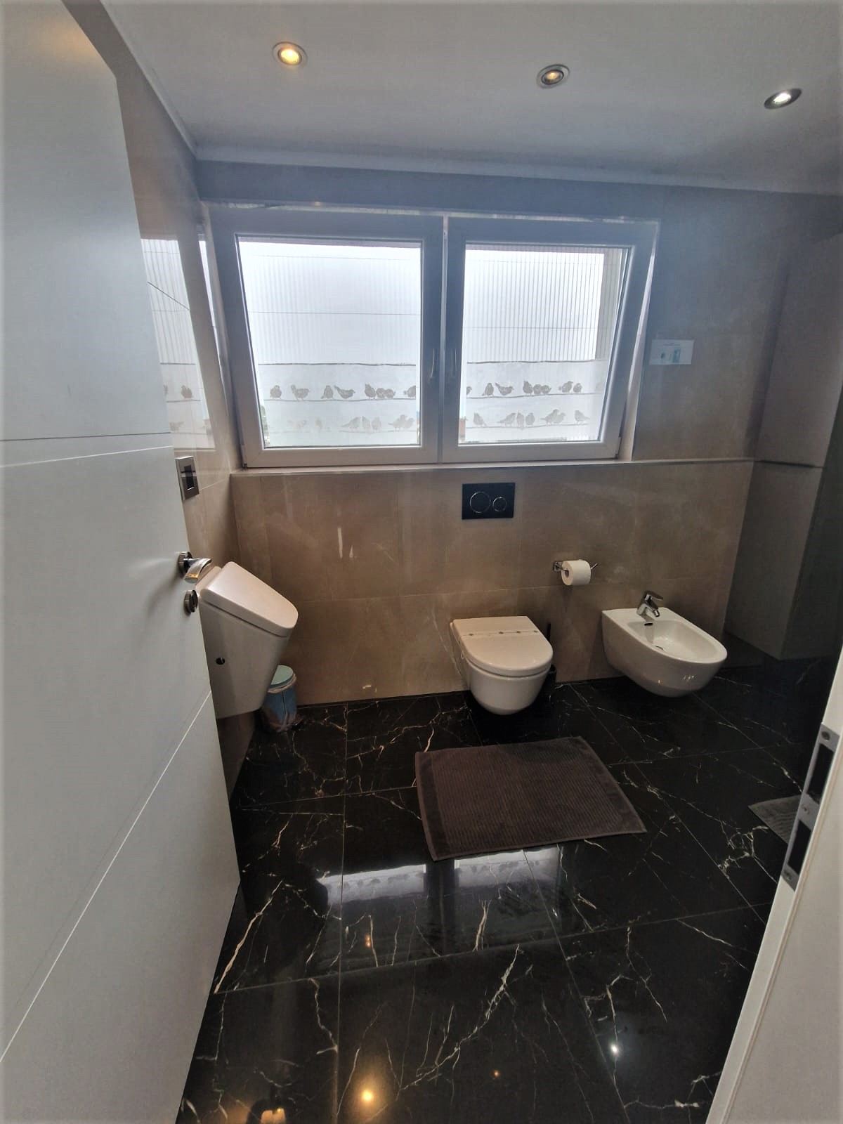 Bathroom corner with a wall-hung toilet and bidet against cream stone tiling below a frosted window, a dark flush plate, and the polished black marble floor running out to the right