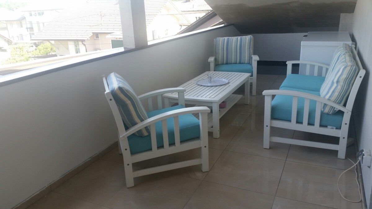 A sheltered rooftop terrace with three white wooden armchairs with turquoise cushions around a low white table, tiled floor, and a view over neighbouring tiled roofs