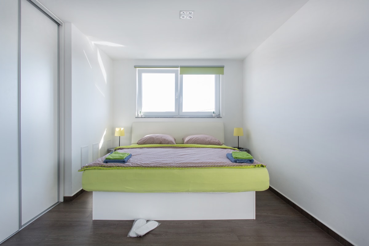 White bedroom with a low white double bed dressed in lime green and mauve, two small lamps either side, a window with a green blind and a full-height frosted sliding wardrobe along the left wall, on dark wood flooring