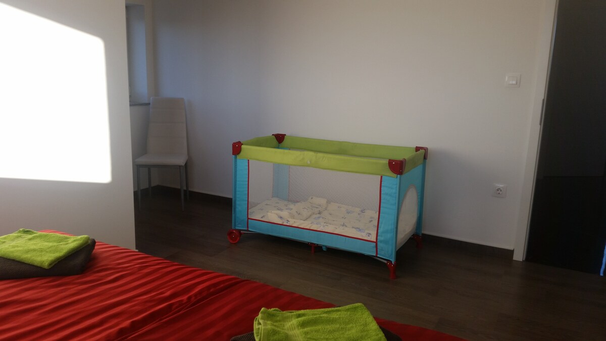 Bedroom with a red-covered bed in the foreground and a blue and green fold-out travel cot made up with bedding standing against the far wall on dark wood flooring