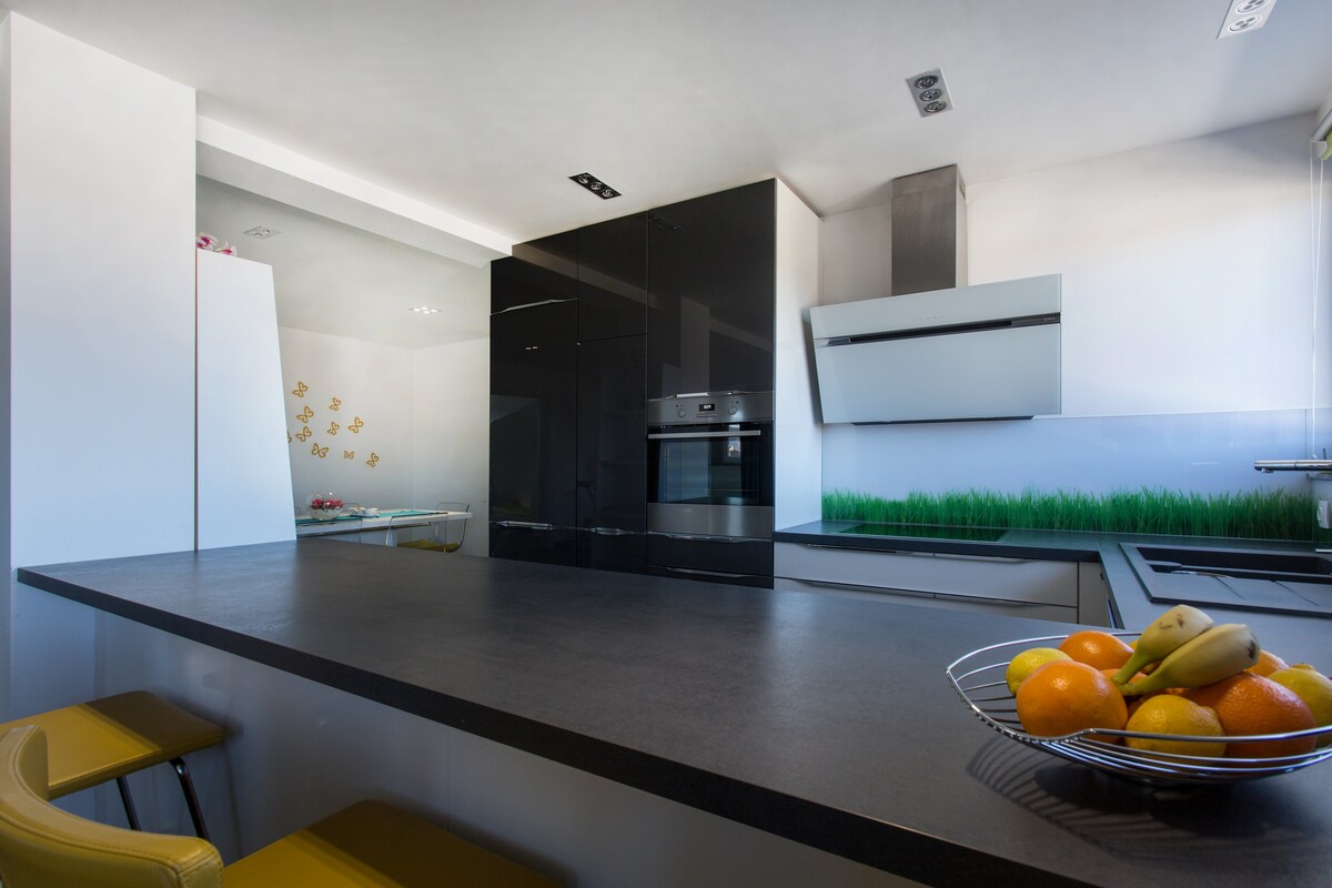 Modern kitchen with a tall black gloss column housing a stainless built-in oven, white gloss base units, a white angled extractor hood, a dark breakfast bar with a bowl of fruit, yellow bar stools and a printed green grass glass splashback
