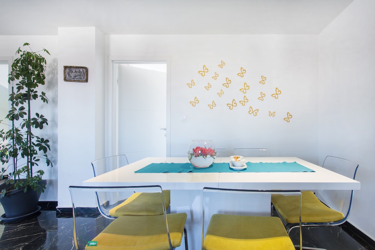 A white gloss dining table with transparent acrylic chairs and yellow seat cushions, a glass bowl of roses on a turquoise runner, and a scatter of gold butterfly decorations on the white wall behind